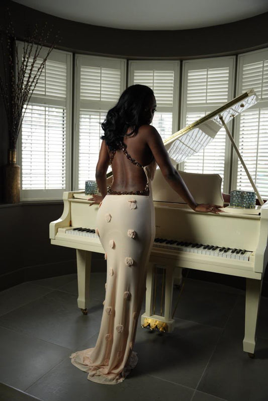 Woman in a floor-length cream AMARAÉ gown with open back, chain straps and 3D rose details, standing at a cream grand piano in soft window light