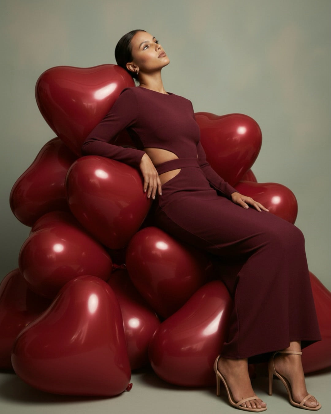 Woman in a long sleeve burgundy AMARAÉ maxi dress with side waist cut-outs and a fitted skirt, seated on a pile of glossy red heart balloons against a sage backdrop, styled with nude strappy heels