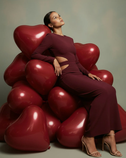 Woman in a long sleeve burgundy AMARAÉ maxi dress with side waist cut-outs and a fitted skirt, seated on a pile of glossy red heart balloons against a sage backdrop, styled with nude strappy heels