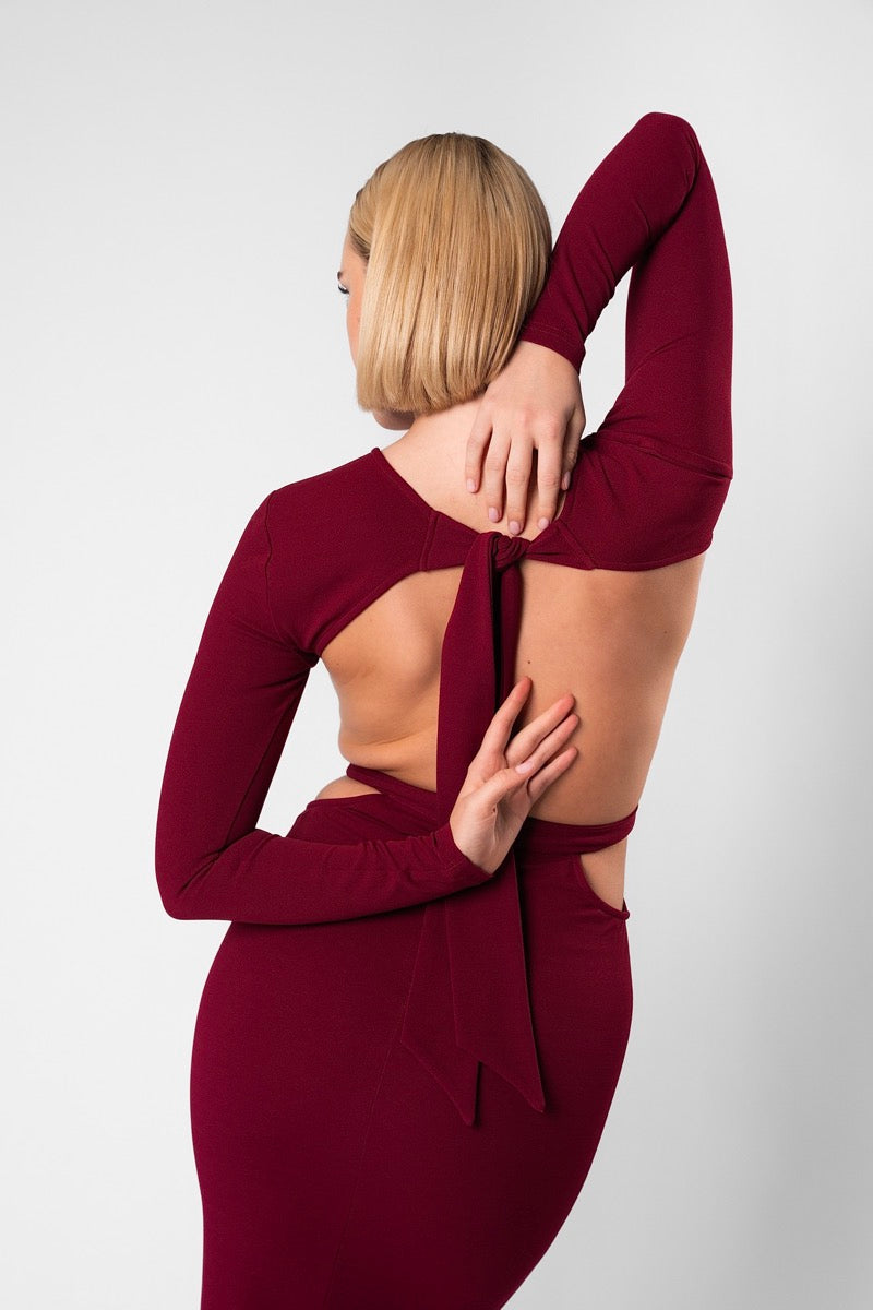 Back view of a woman in the burgundy AMARAÉ Coco De Mer long sleeved dress with an open back, a tie at the shoulders and waist cut outs, against a plain light grey studio backdrop