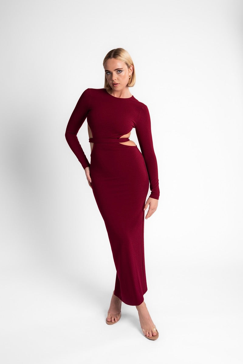 Woman in the burgundy AMARAÉ Coco De Mer long sleeved maxi dress with a crew neckline and cutouts at the waist, against a plain light grey studio backdrop