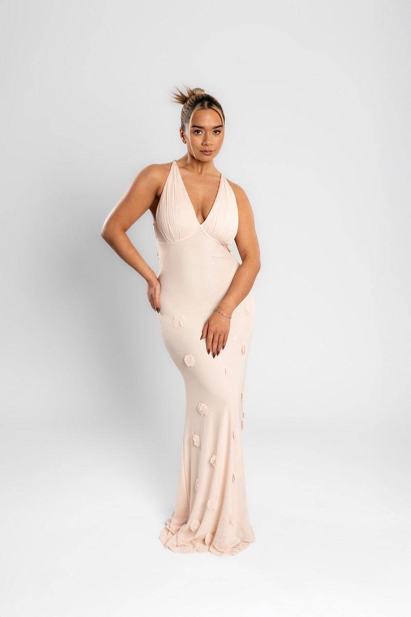 Woman in the blush AMARAÉ Willow maxi dress with a plunging halter V neckline, a seam under the bust and 3D rosettes on a flowing floor length skirt, against a plain light grey studio backdrop